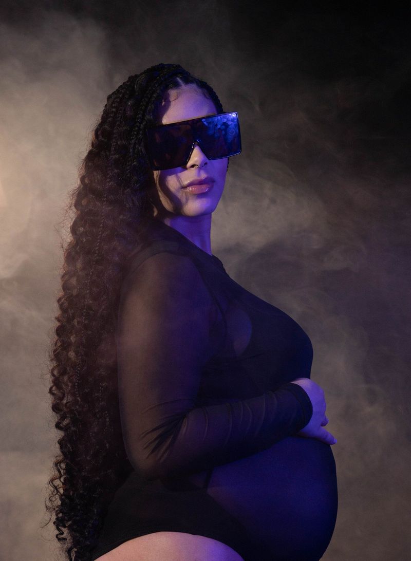 Maternity portrait of a woman in sunglasses with long braids in purple light and haze