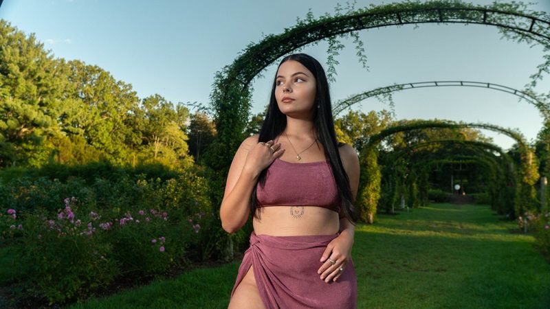 A woman with long dark hair in a mauve two-piece standing beneath vine-covered garden arches at golden hour