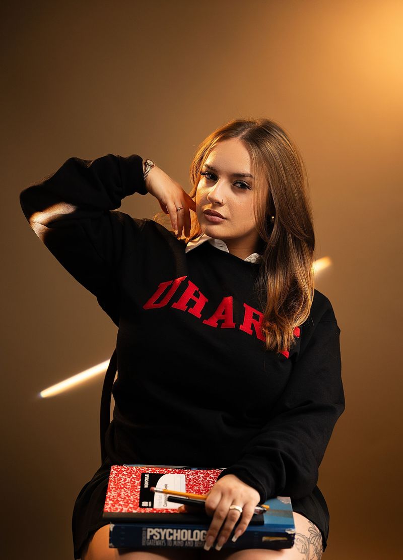 A senior portrait of a young woman in a black sweatshirt with red lettering, holding notebooks, in warm studio light