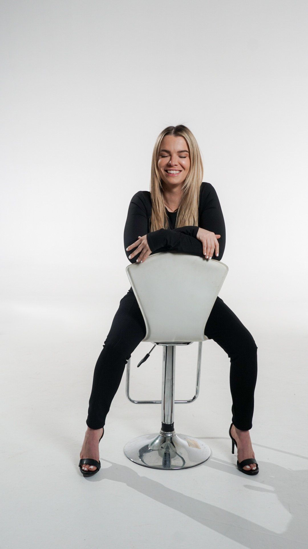 A smiling woman in black leaning on the back of a white stool against a seamless white studio backdrop