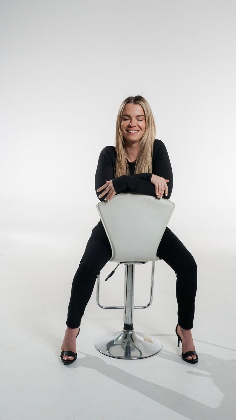 A smiling woman in black leaning on the back of a white stool against a seamless white studio backdrop