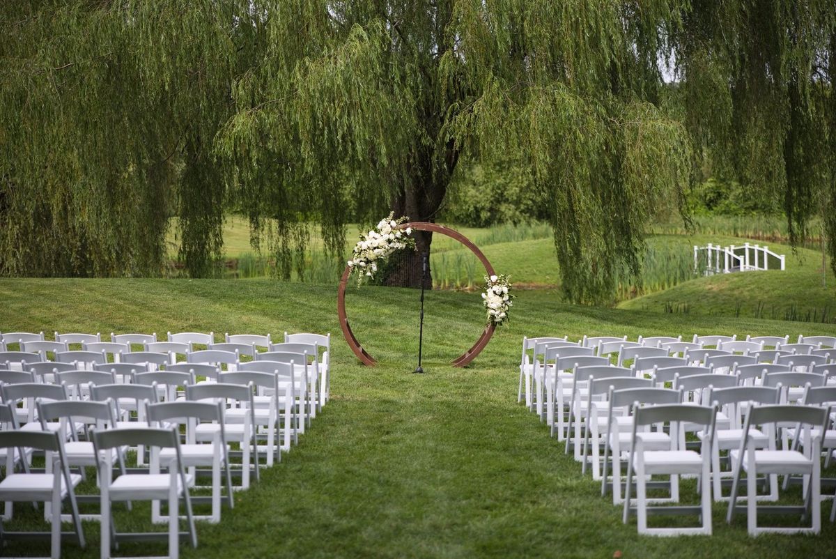 White ceremony chairs lead to a circular floral arch beneath the willows