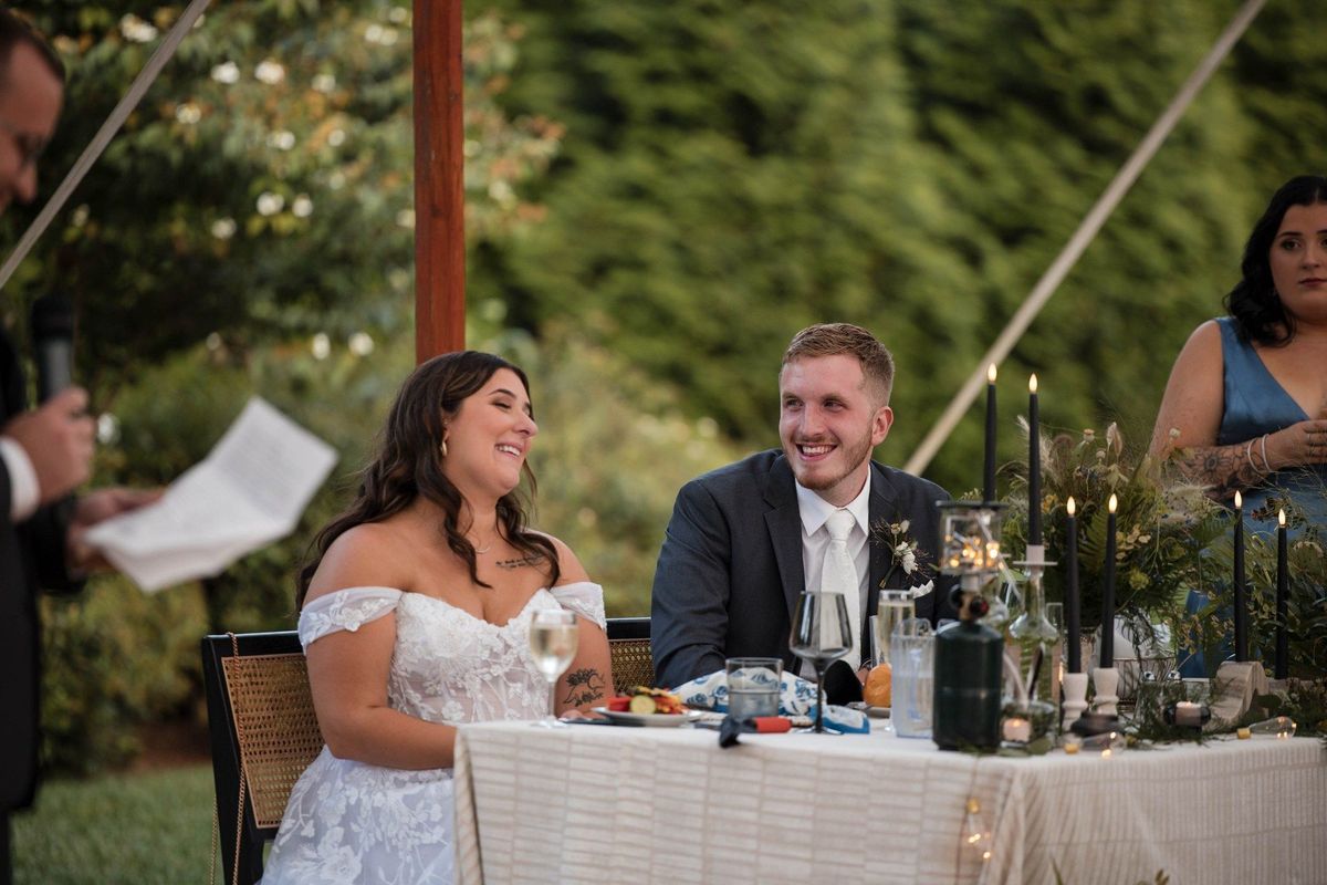 Sophia and Tommy smile together at their reception table while listening to speeches