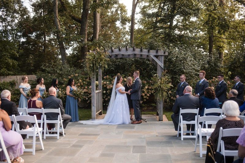 A wide view shows Sophia and Tommy beneath the arbor with seated ceremony guests