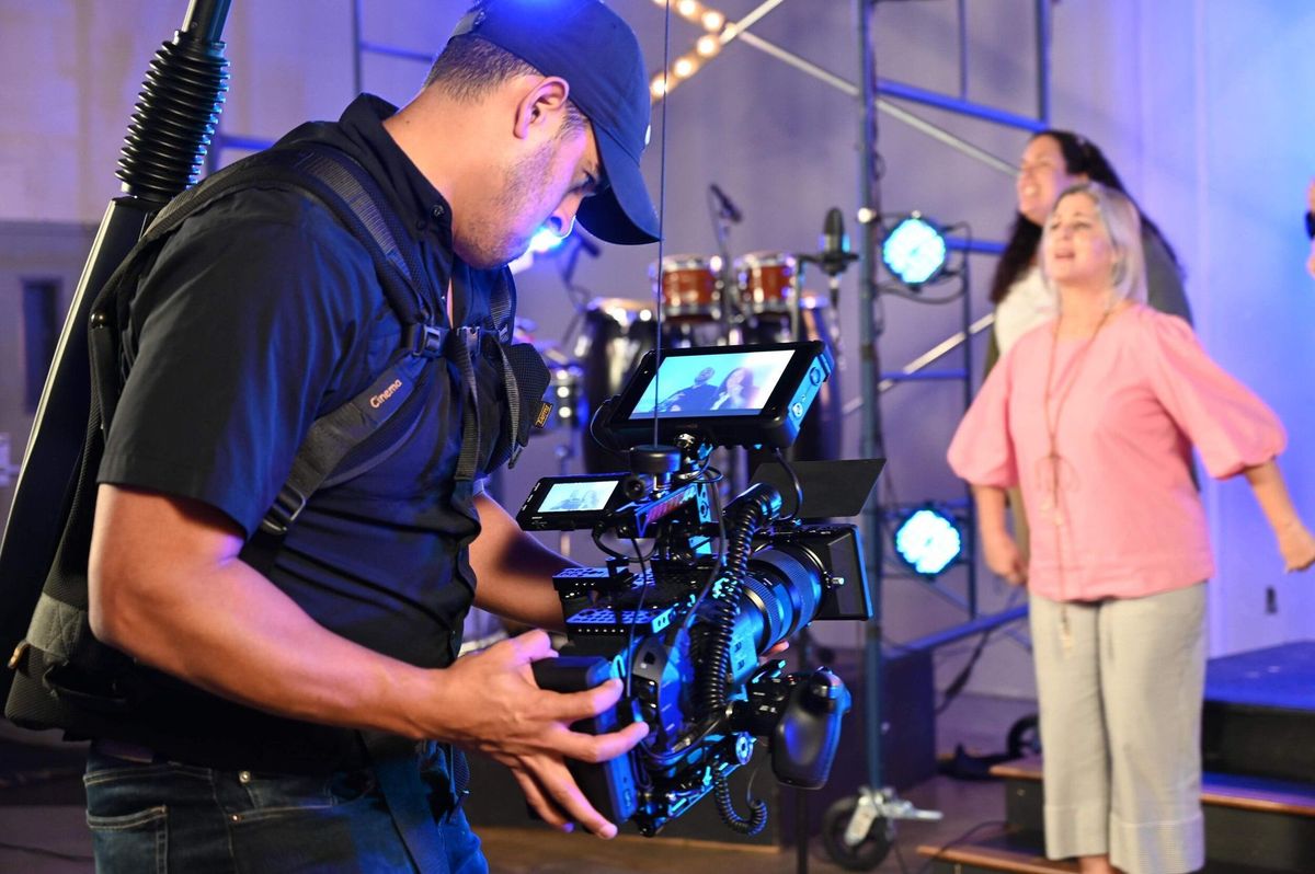 A camera operator in a cap videos two women singing on a stage lit in blue and purple