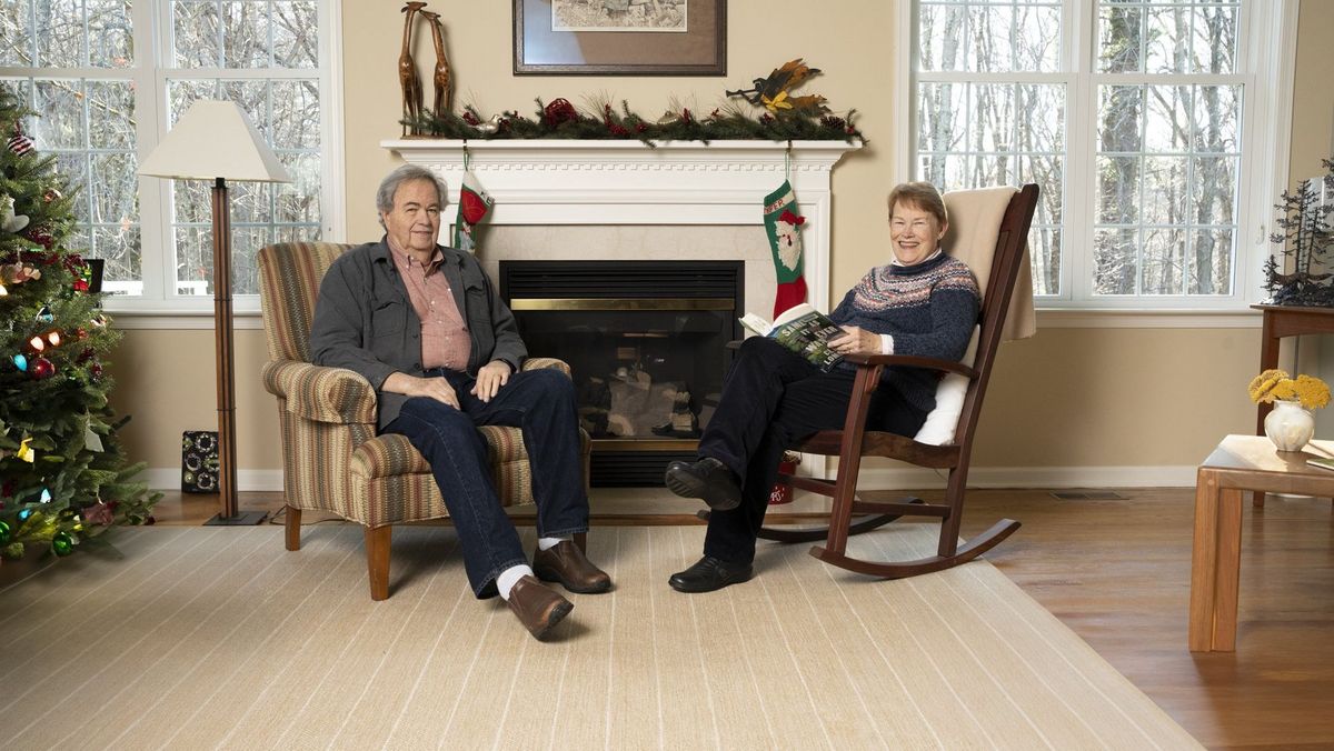 Two older adults seated in armchairs beside a fireplace and large windows
