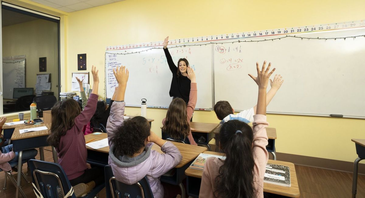 Students raising their hands while a teacher points to a classroom whiteboard