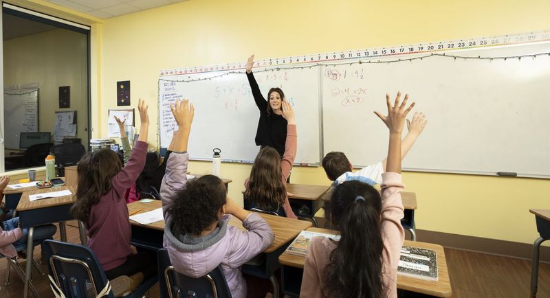 Students raising their hands while a teacher points to a classroom whiteboard
