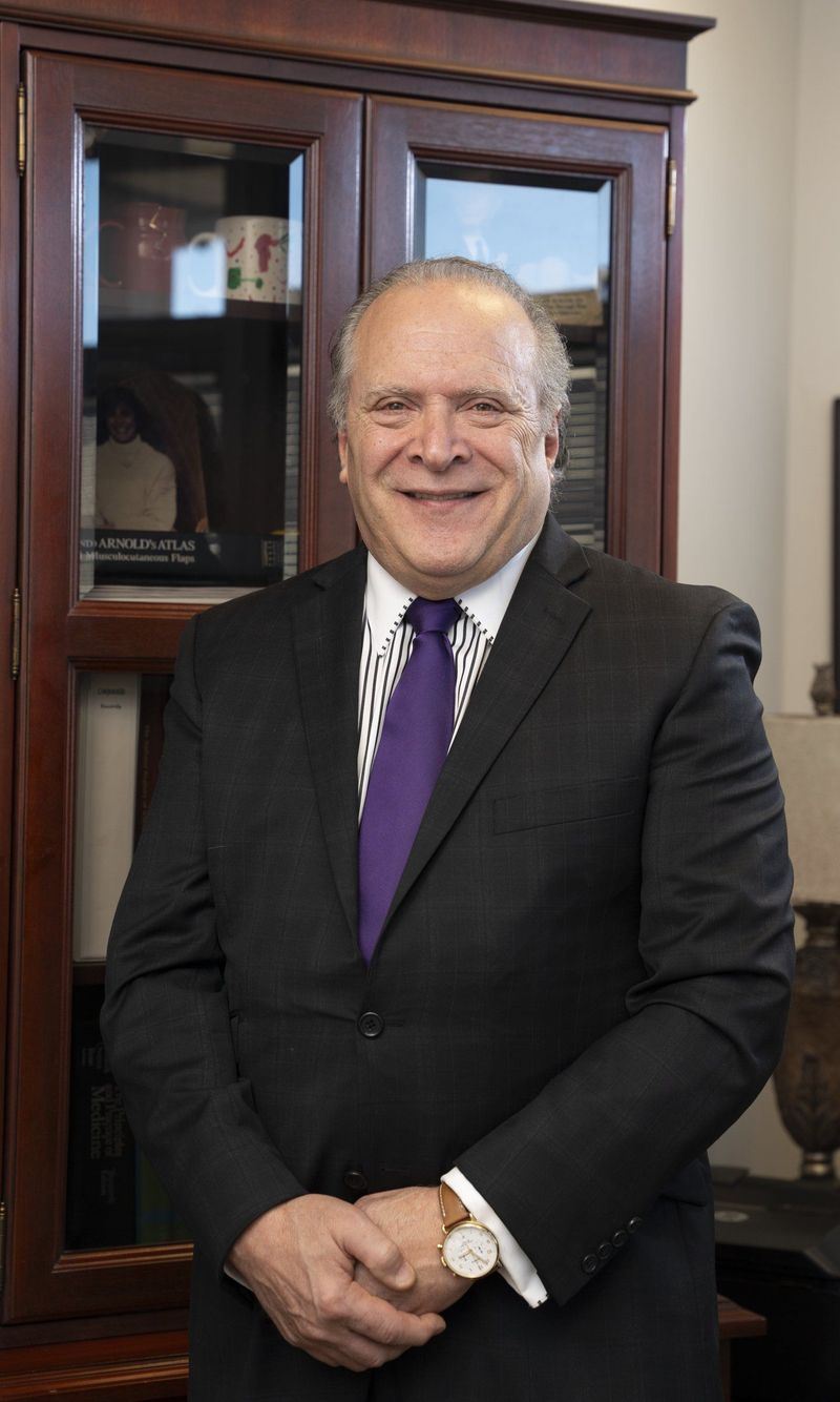 Man in a dark suit and purple tie posed beside a wooden cabinet