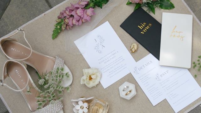 Flat lay of wedding invitations, vow books, a ring, decorated shoes and flowers from the opening of Adam and Jess's wedding film