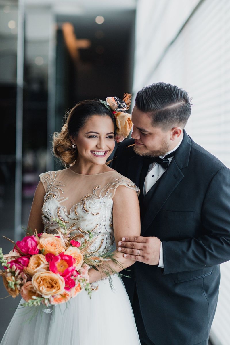 A smiling bride in a beaded gown holding a pink and peach bouquet while her groom in a tuxedo leans in beside her