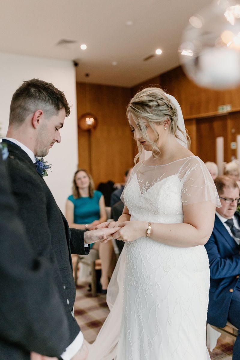 A groom in a kilt jacket placing a ring on his bride's finger during an indoor ceremony as guests watch