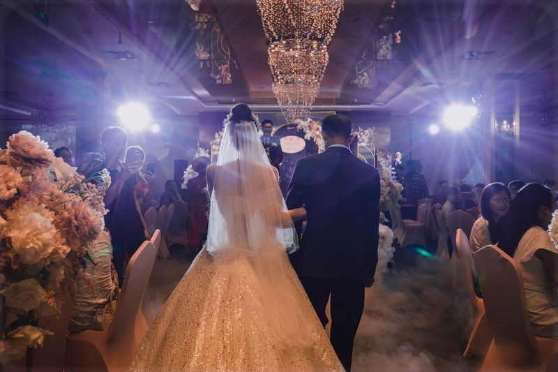 A bride in a veil and a groom in a navy suit walking arm in arm through a ballroom lit by stage lights and a chandelier, guests photographing them