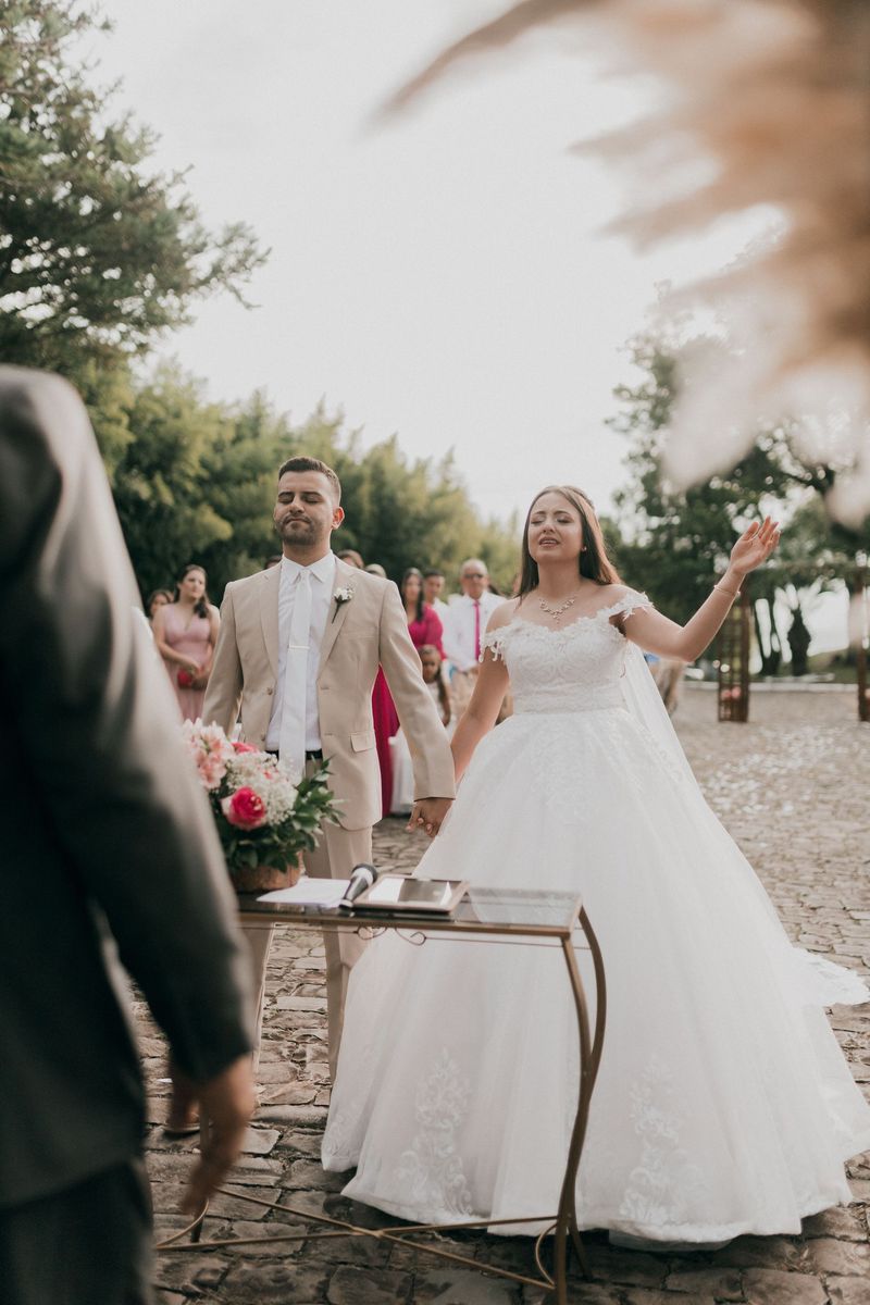 A bride and groom in a beige suit holding hands at an outdoor ceremony with their eyes closed, guests behind them
