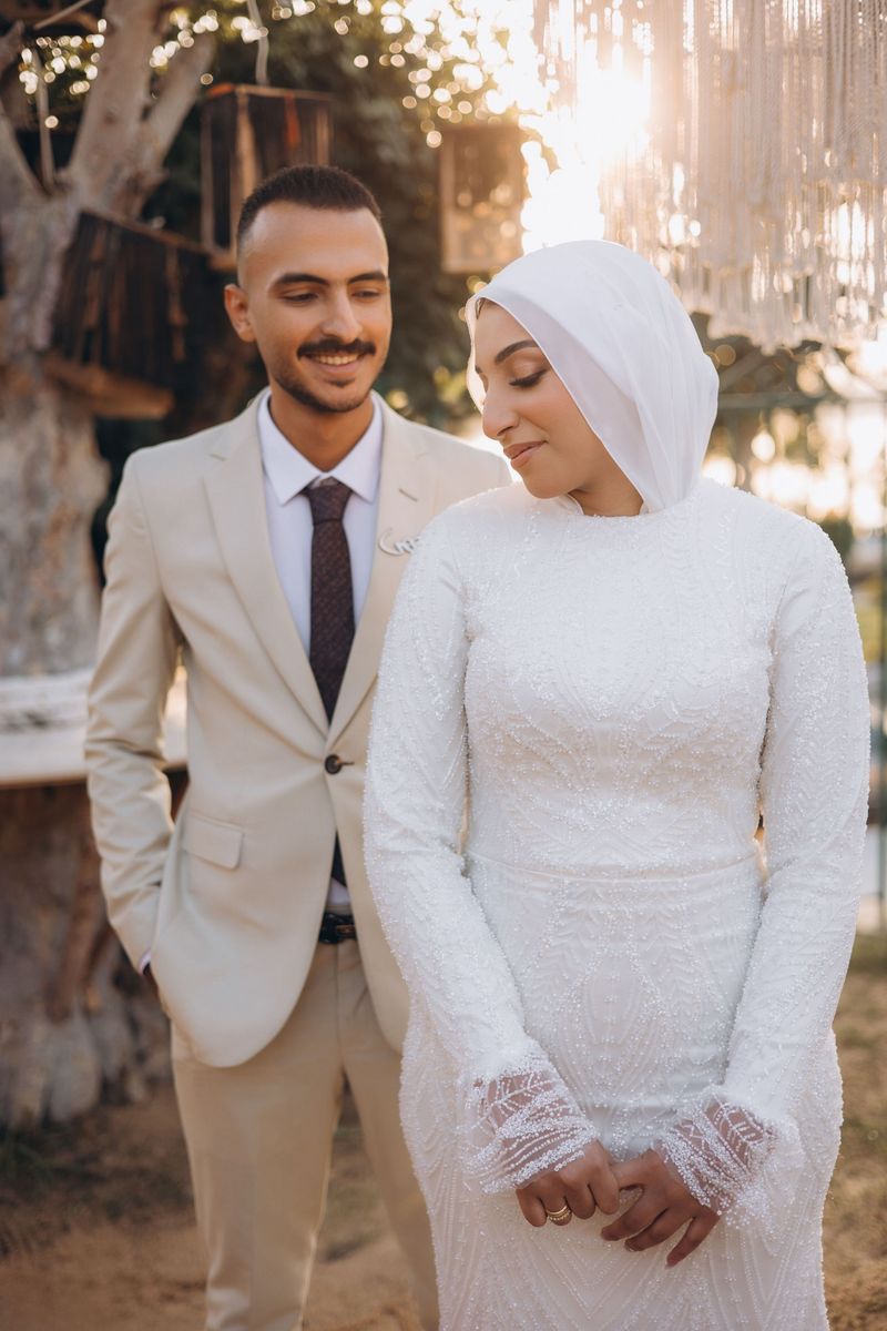 A bride in a beaded white gown and headscarf beside a groom in a beige suit and patterned tie