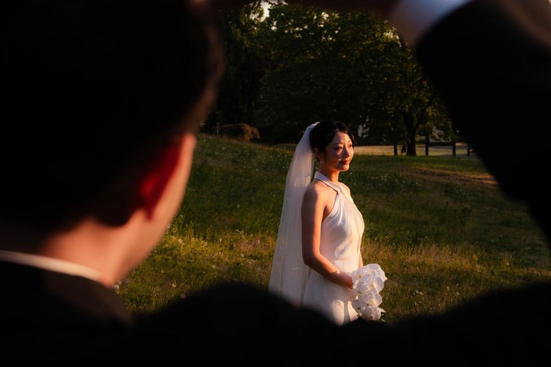 A bride in a veil holding a white orchid bouquet on a grassy lawn in warm evening light, seen between two blurred guests