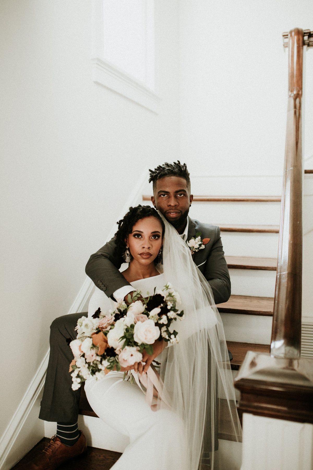 A bride in a long veil holding a blush bouquet, seated on a wooden staircase in front of her groom in a gray suit