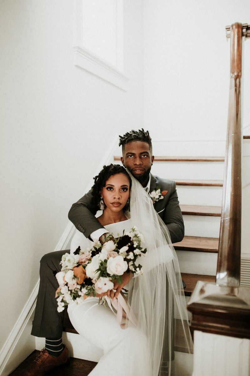A bride in a long veil holding a blush bouquet, seated on a wooden staircase in front of her groom in a gray suit