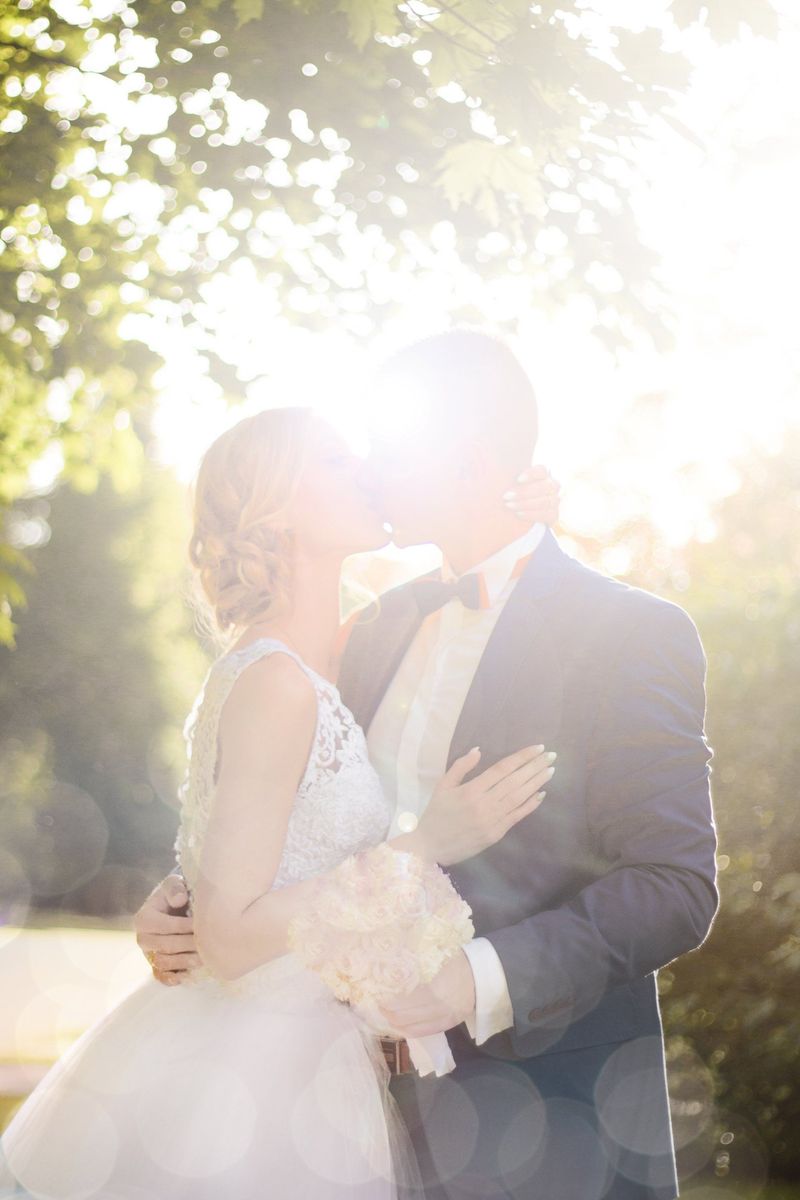 A bride and groom kissing in bright sunlight beneath leafy trees