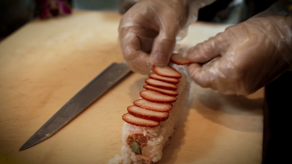 Hands arrange sliced strawberries over a sushi roll