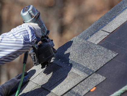 Worker installing dark roof shingles with a nail gun on a sloped roof