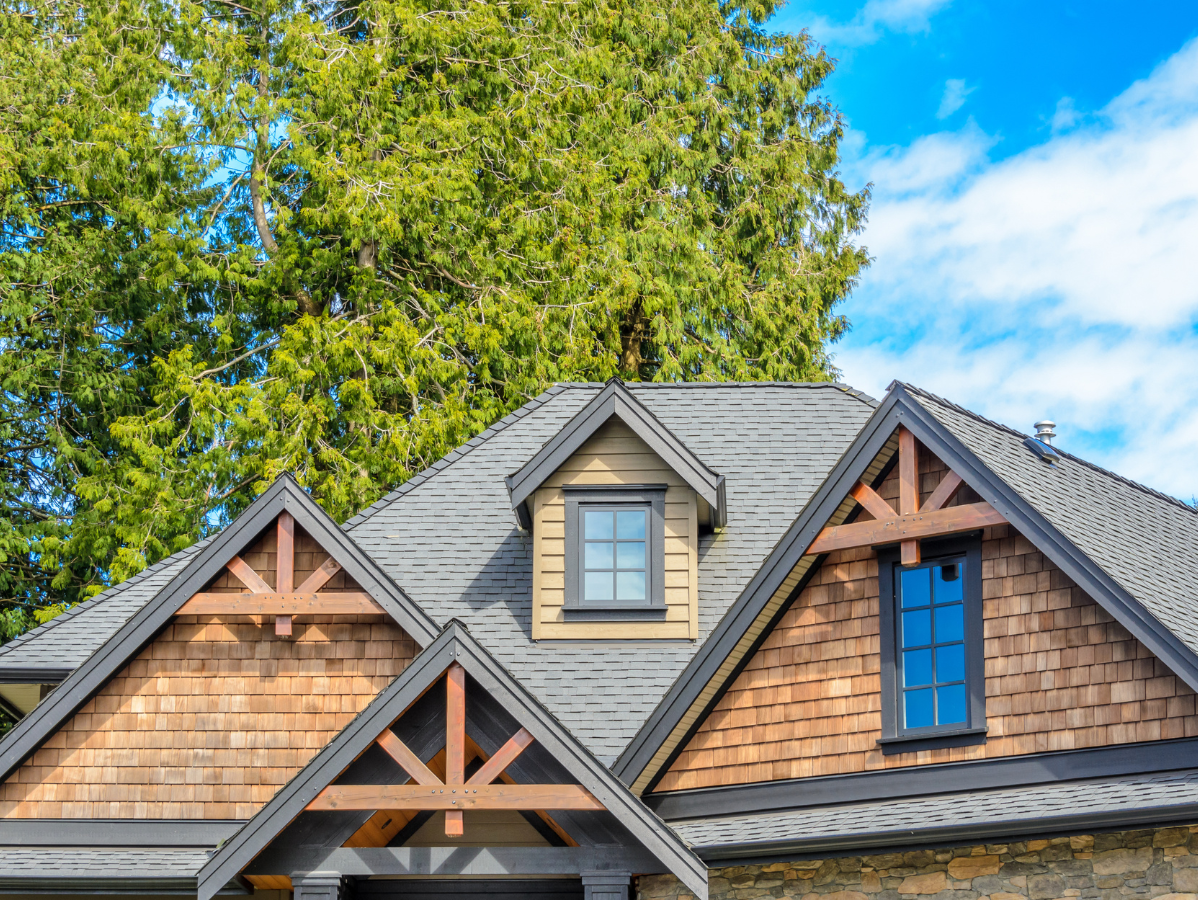 House roof with gray shingles, wooden gables, and blue windows under a leafy green tree and blue sky