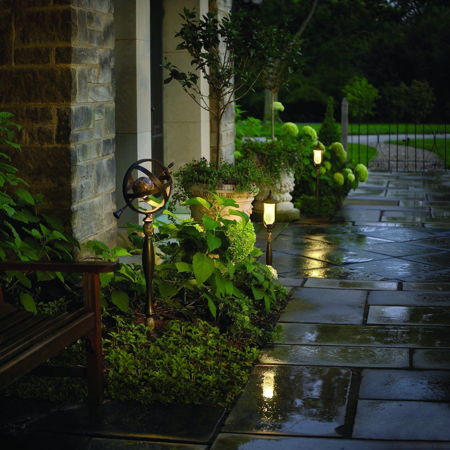 Pathway lit by garden lights, alongside lush greenery, and a stone facade.