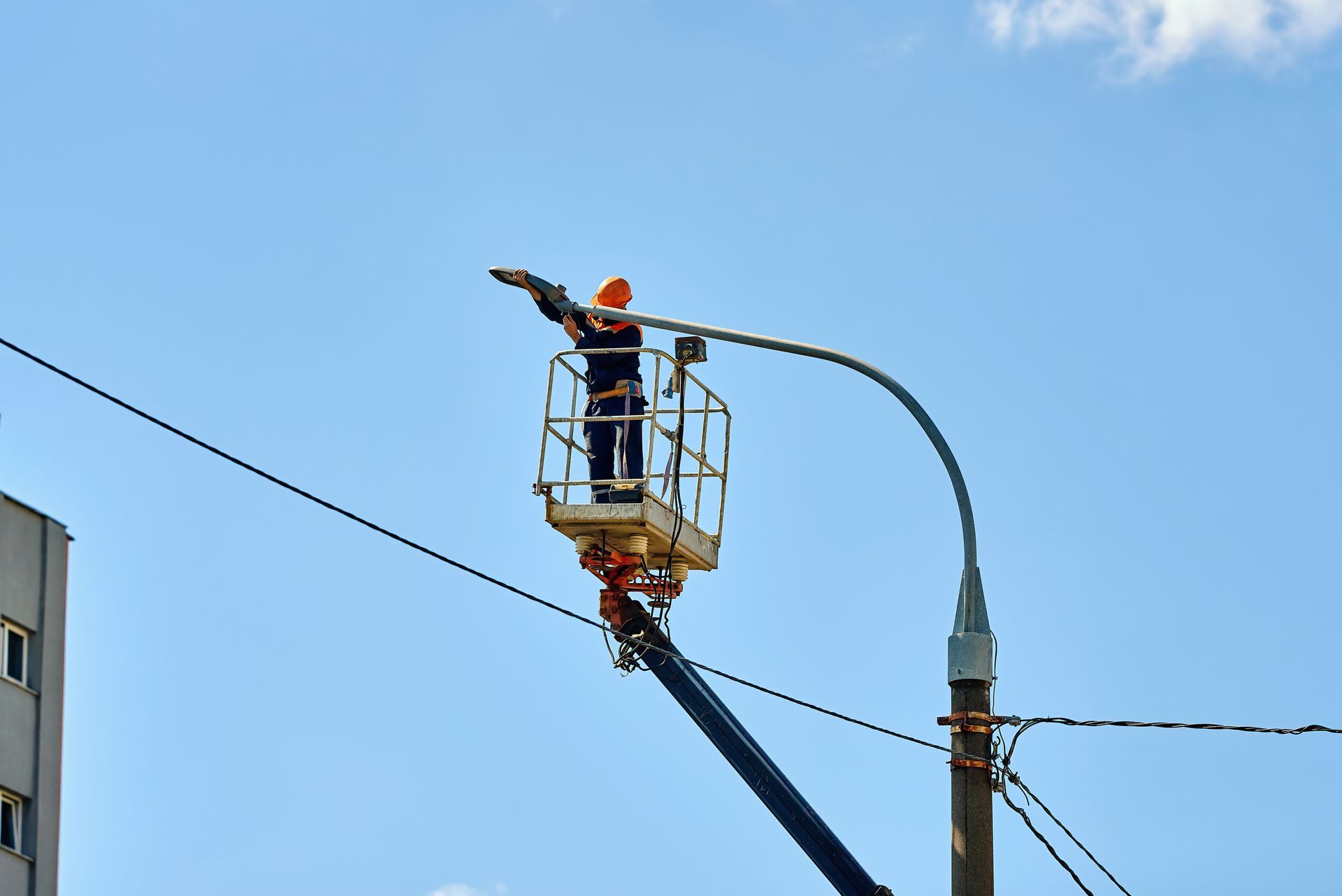 Worker in an orange hard hat on a lift fixing a street light against a blue sky.