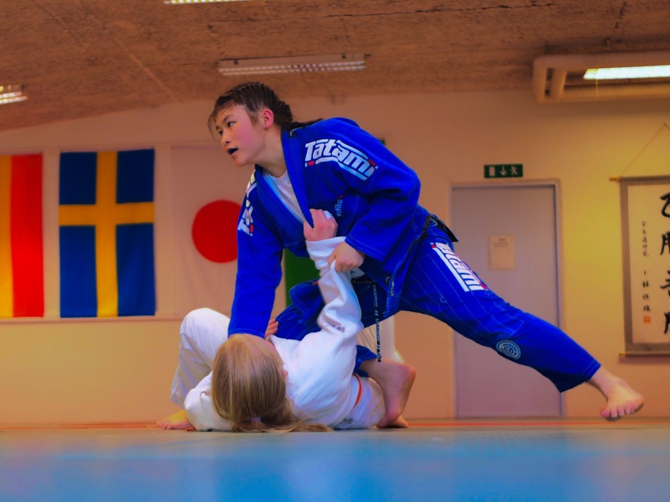 Två personer utövar Jiu-Jitsu. En i blå gi, stående, greppar tag i benet på personen i vit gi, som ligger på golvet.
