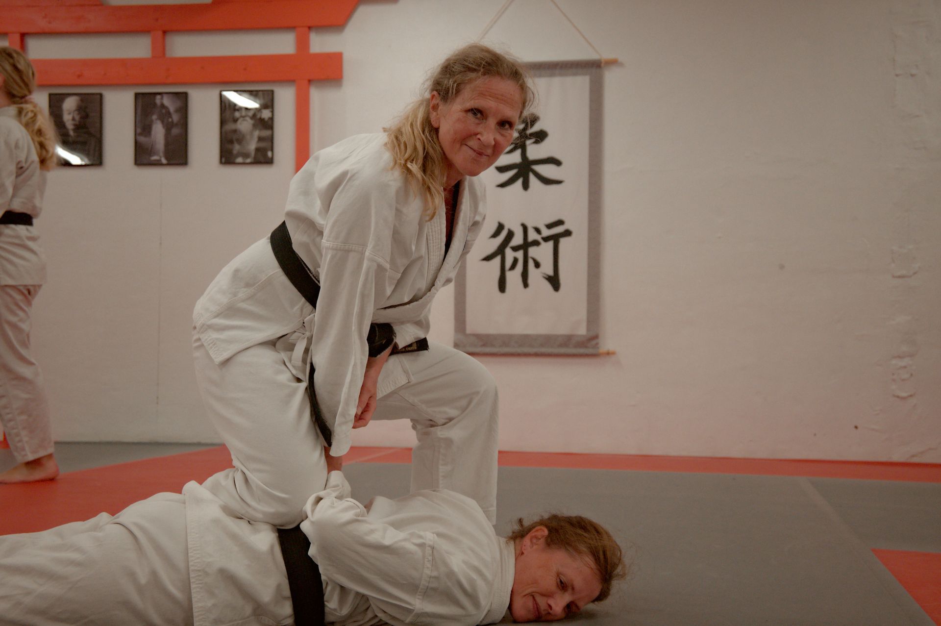 Kvinna i vit gi knäböjer på en annan i en judodojo, leende. Japansk kanji-rulle i bakgrunden.