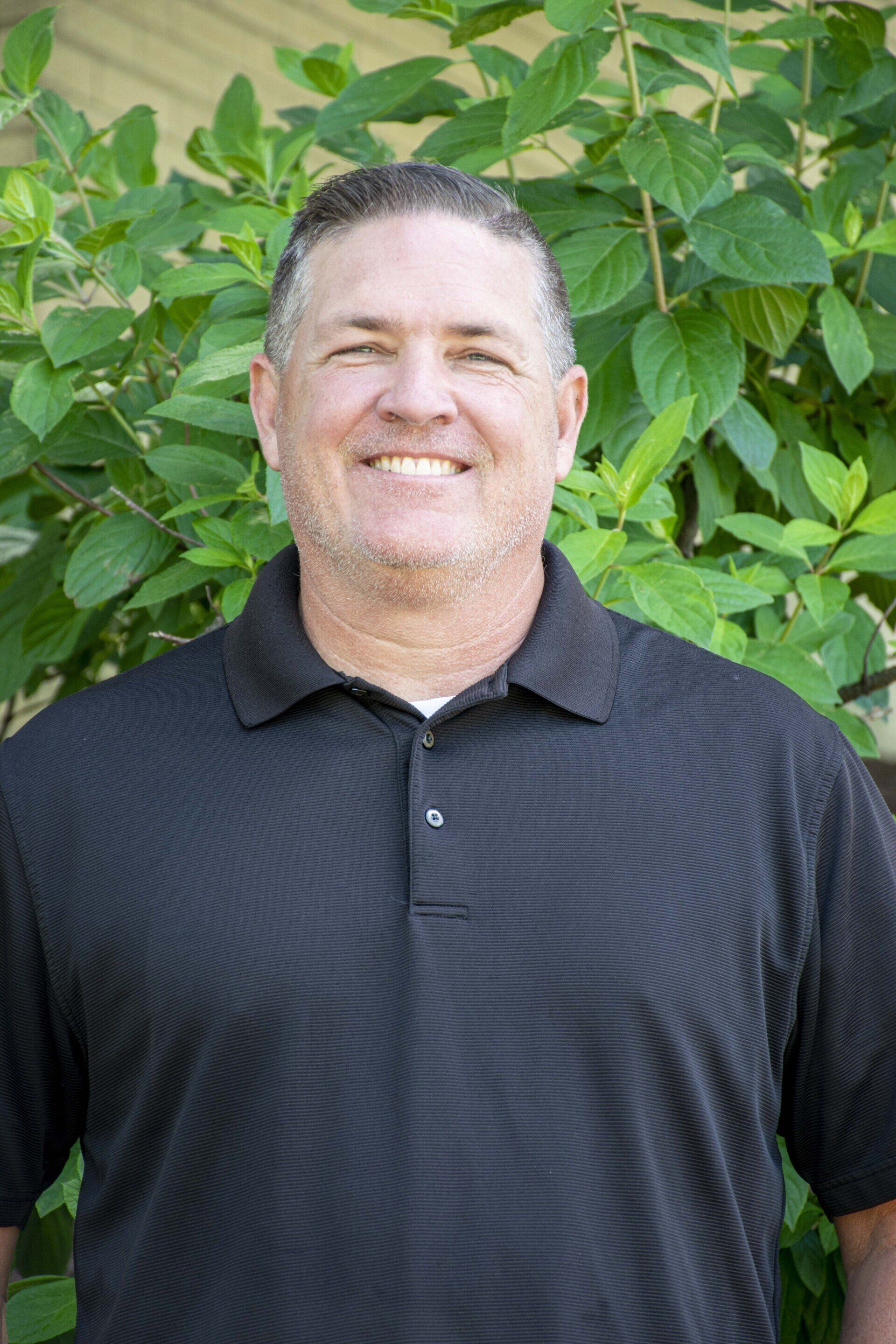 Man smiling, wearing black polo, standing in front of green foliage.