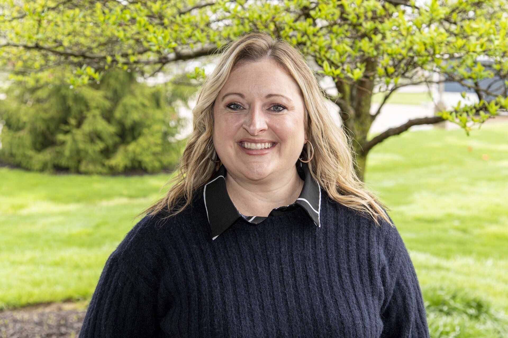 Woman with blonde hair smiles outdoors in front of a tree; she wears a dark sweater.