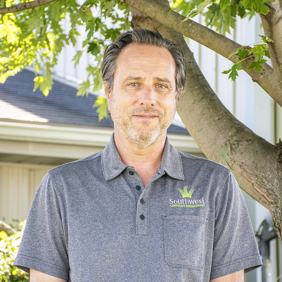 Man in gray polo shirt with logo smiles in front of tree and building.