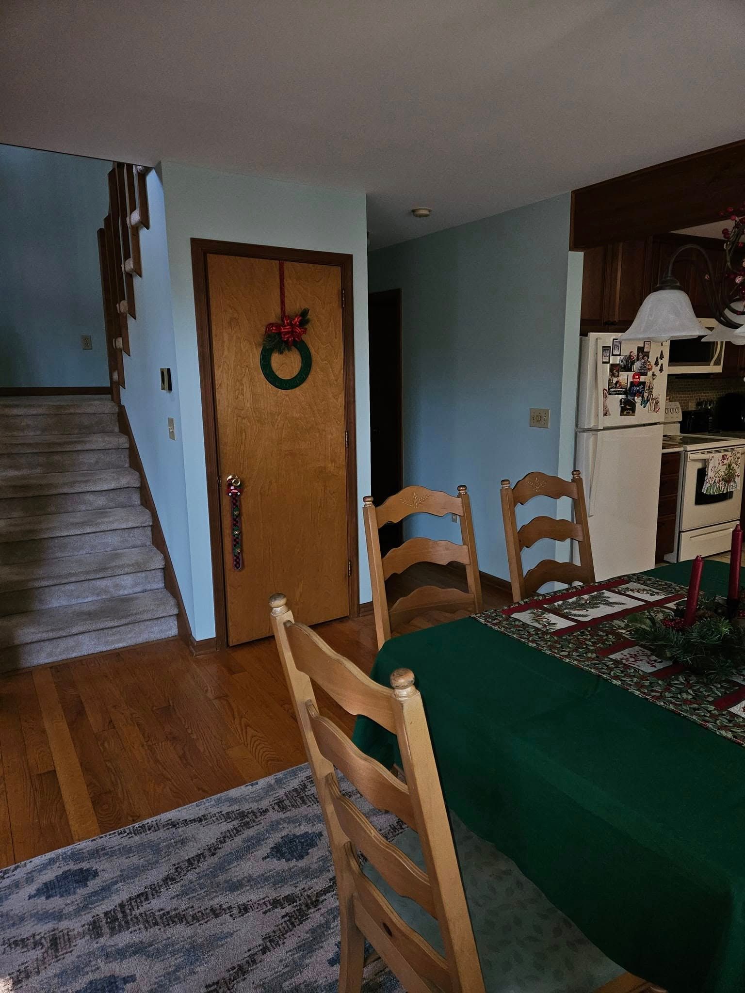 Dining room with green tablecloth, wooden chairs, staircase, and kitchen area in the background