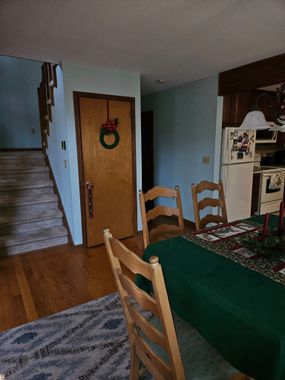 Dining room with green tablecloth, wooden chairs, staircase, and kitchen area in the background