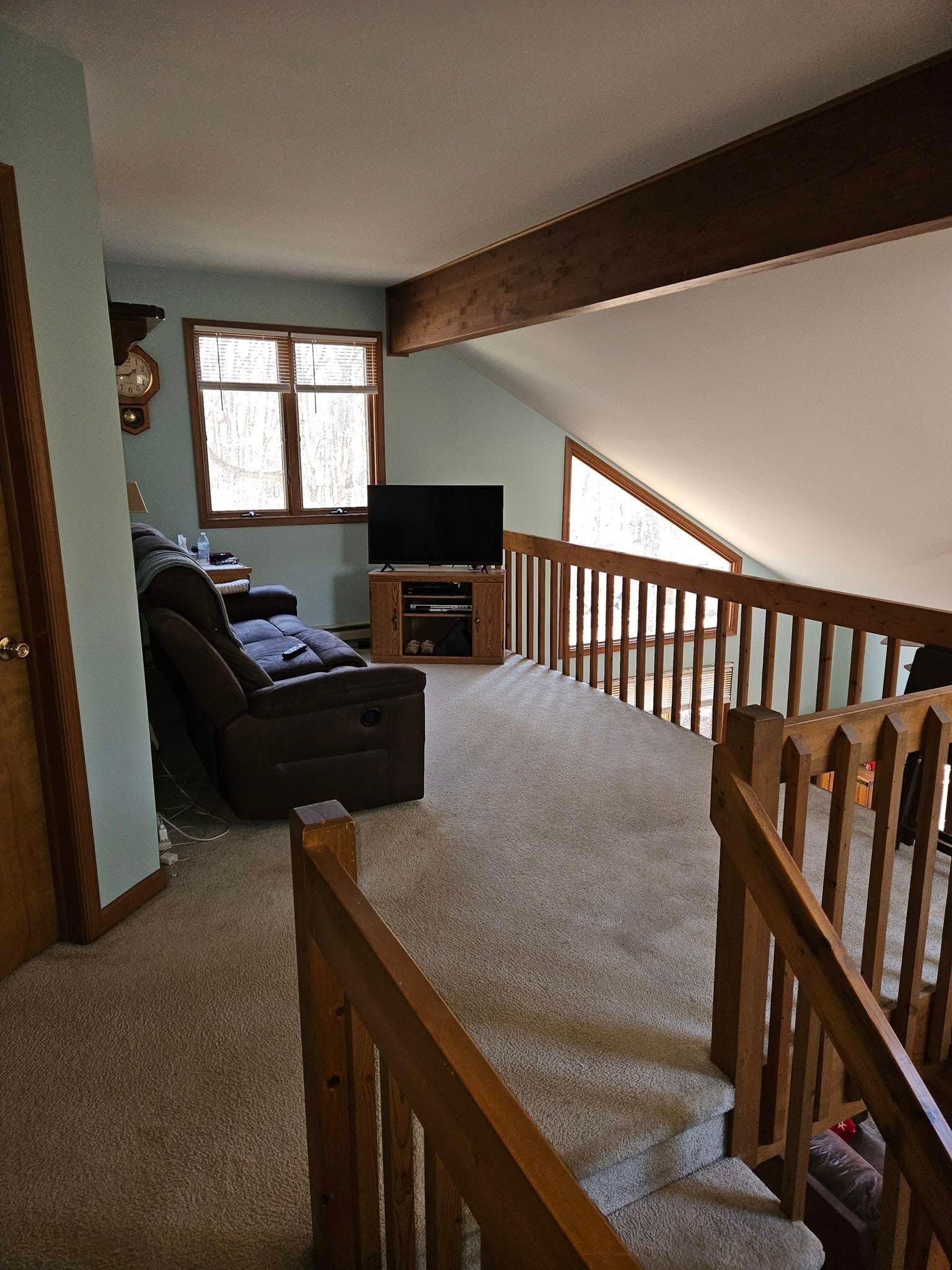 Carpeted loft landing with brown sofa, TV, and wooden railing by a window
