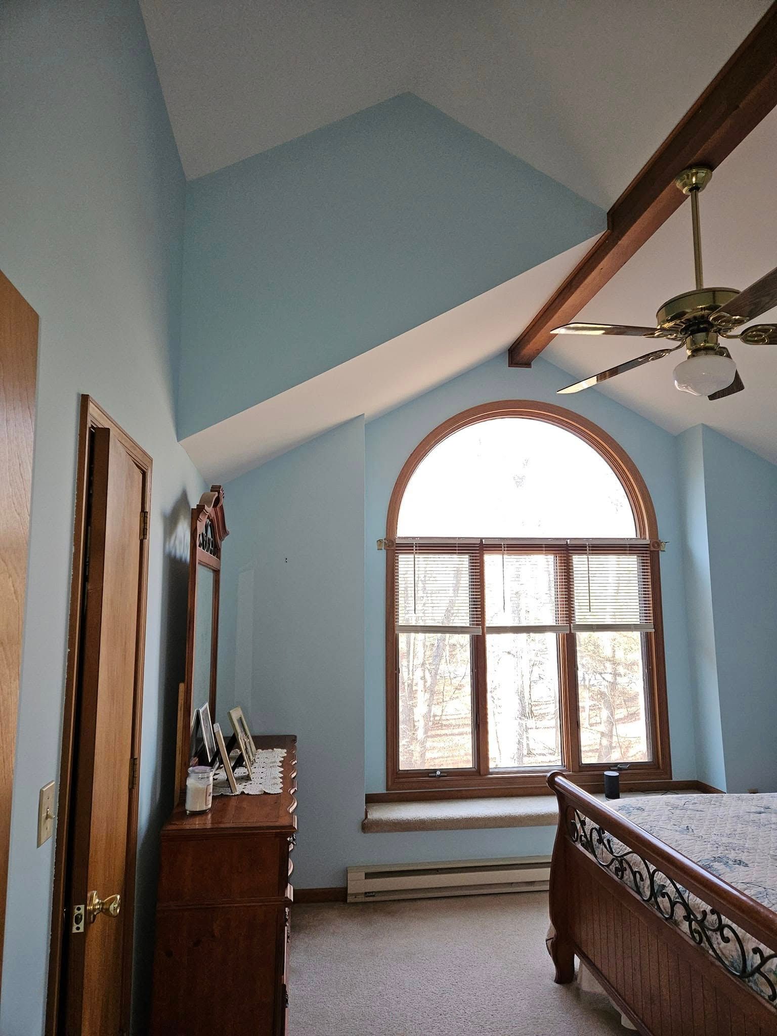 Bedroom with light blue walls, arched window, wooden dresser, and a bed under a ceiling fan.
