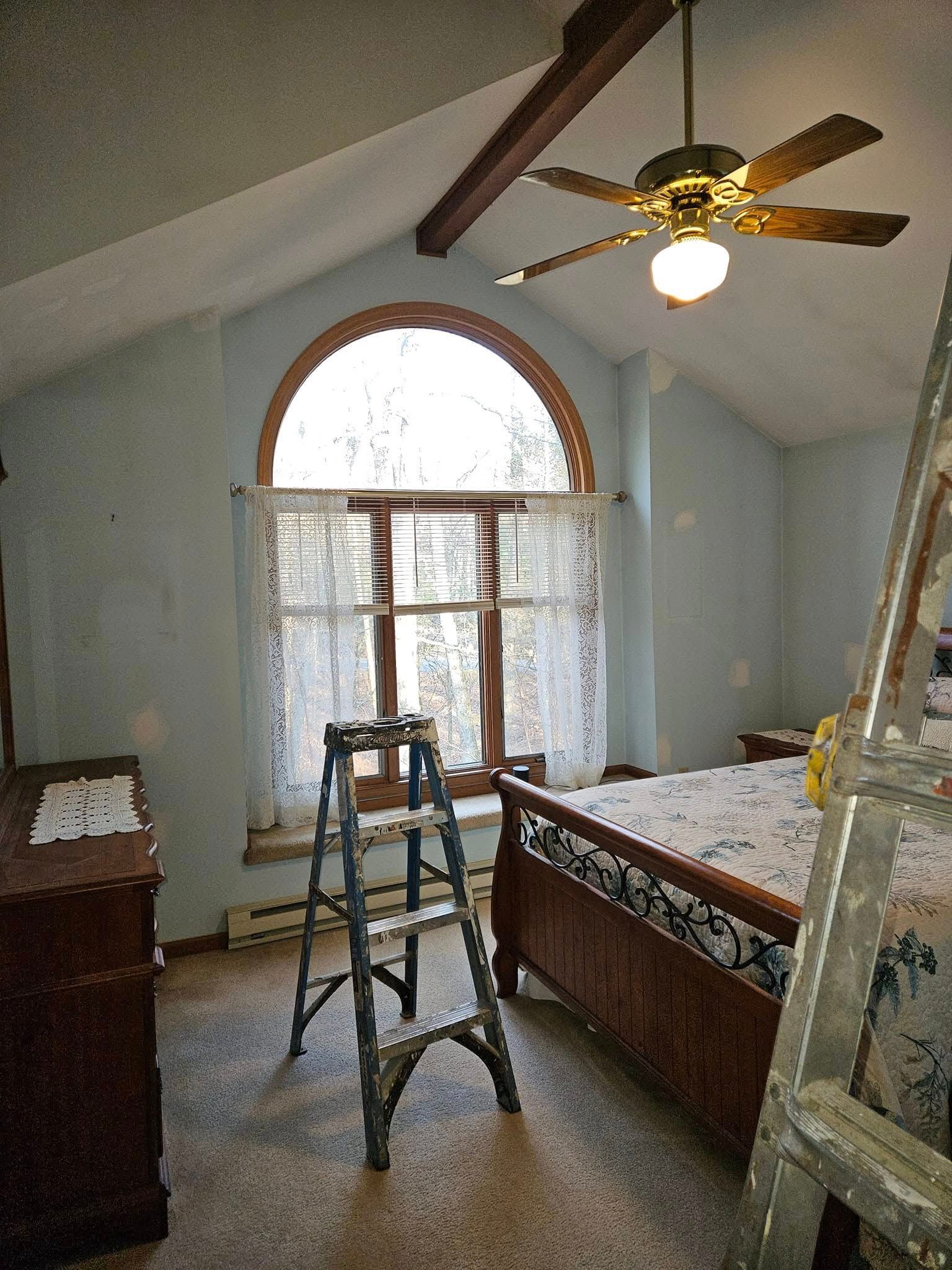 Bedroom under renovation with ladder, ceiling fan, and arched window with sheer curtains