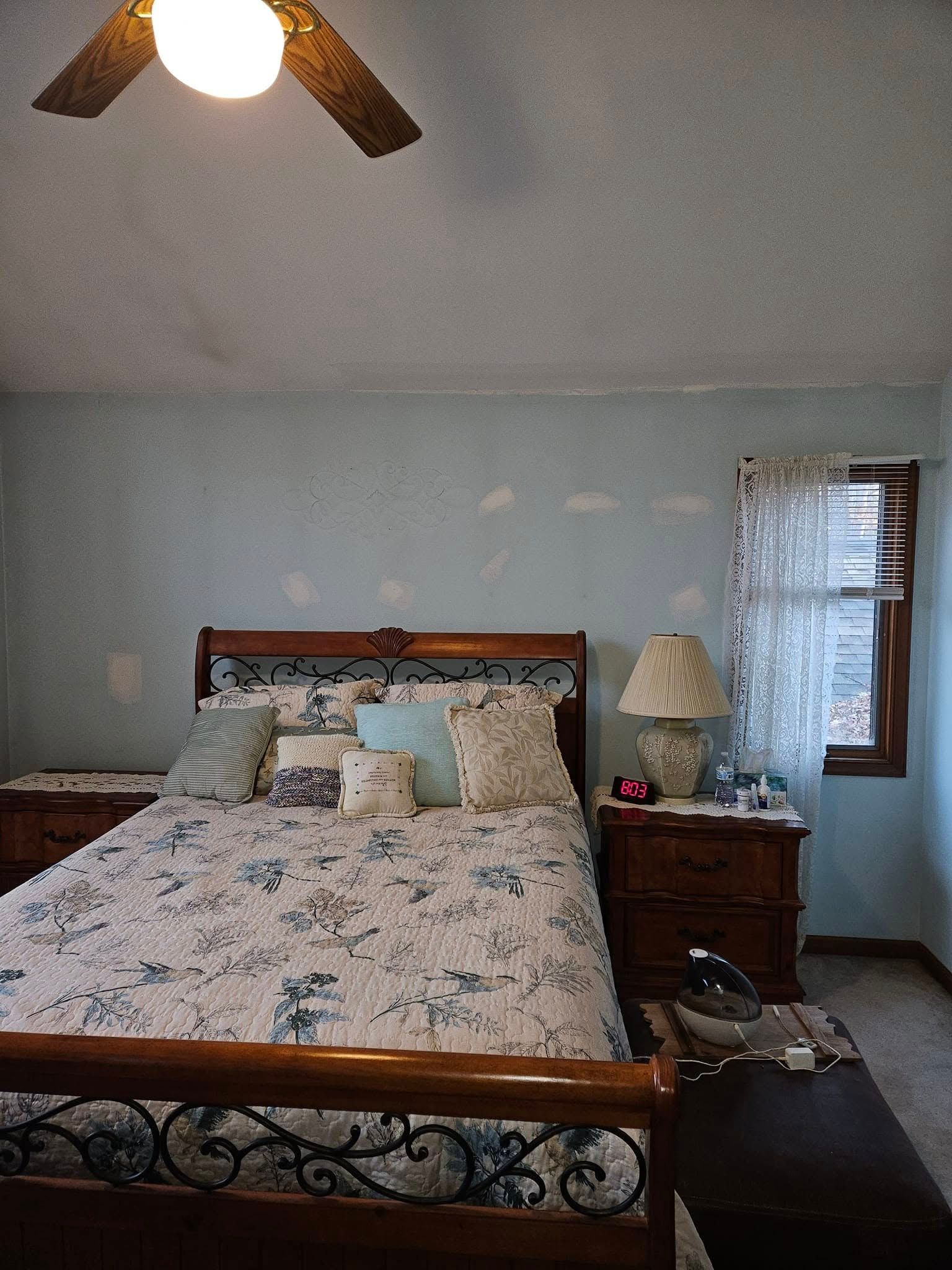 Bedroom with floral bedding, wooden nightstand, ceiling fan, and window with lace curtain.