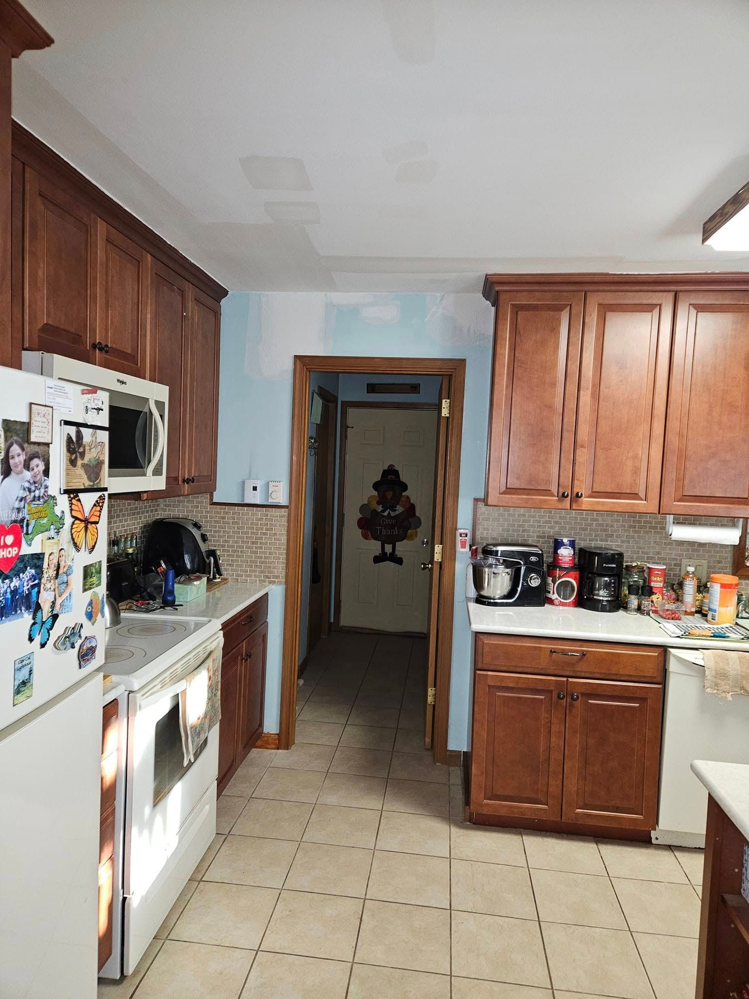 Narrow kitchen with wood cabinets, white appliances, tiled floor, and a hallway opening at the back.
