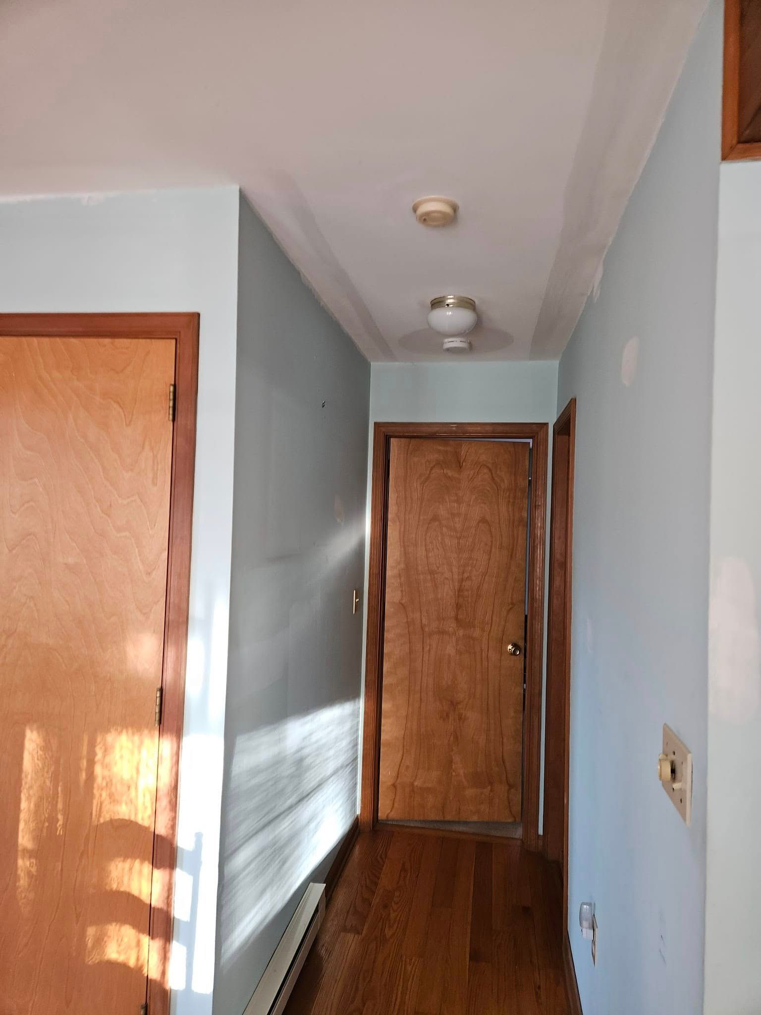 Narrow hallway with light blue walls, wood doors, and sunlight casting shadows on the floor
