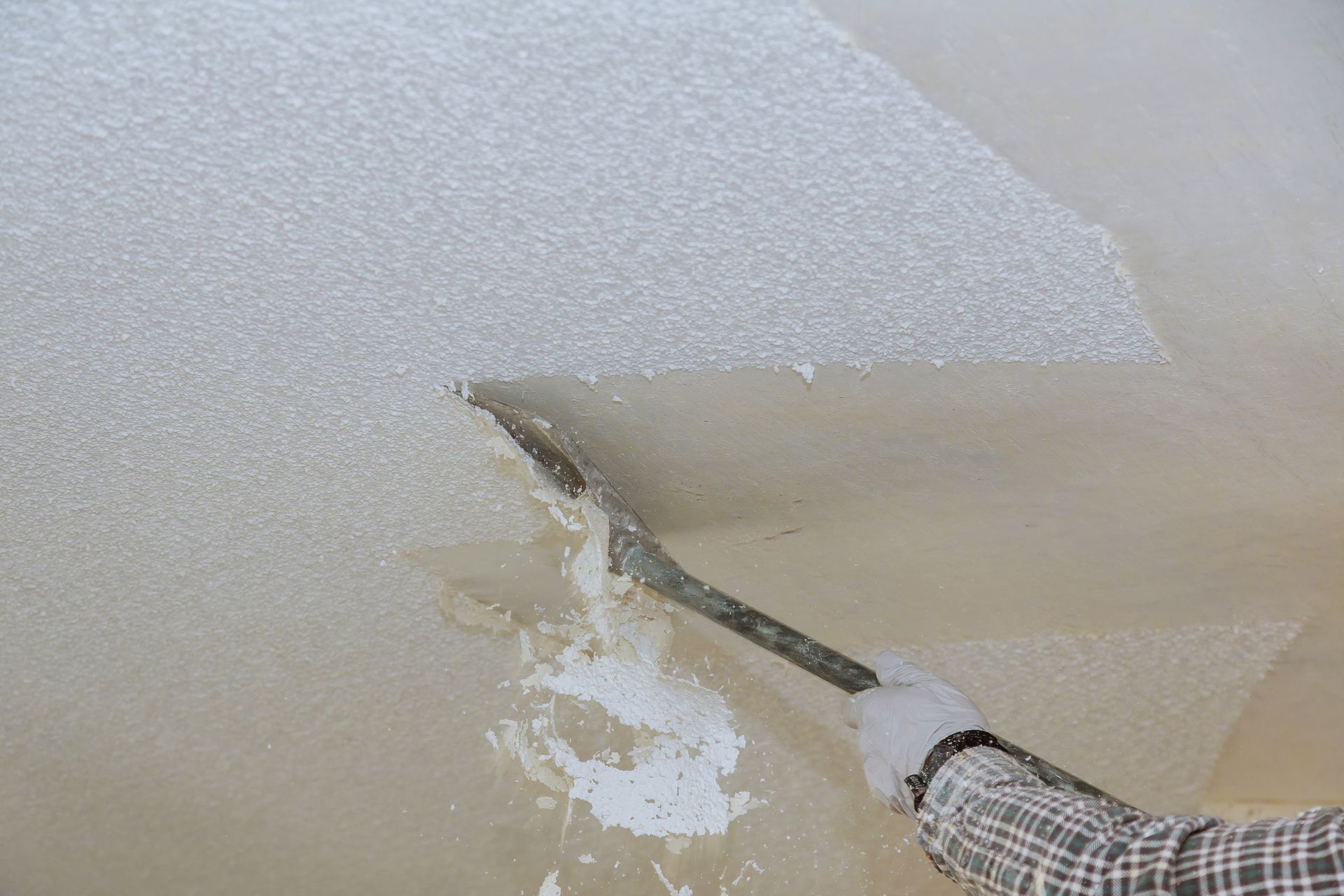 Person scraping peeling ceiling paint with a putty knife, exposing damaged plaster
