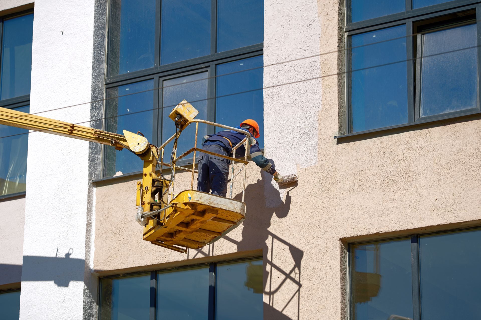 Worker in a yellow cherry picker repairing a building exterior near blue windows.