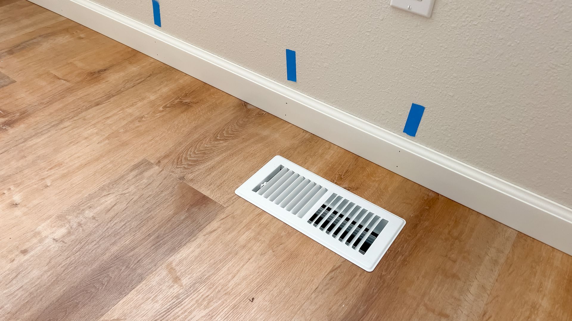 Floor vent in a light wood room beside a white baseboard with blue painter’s tape marks on the wall.