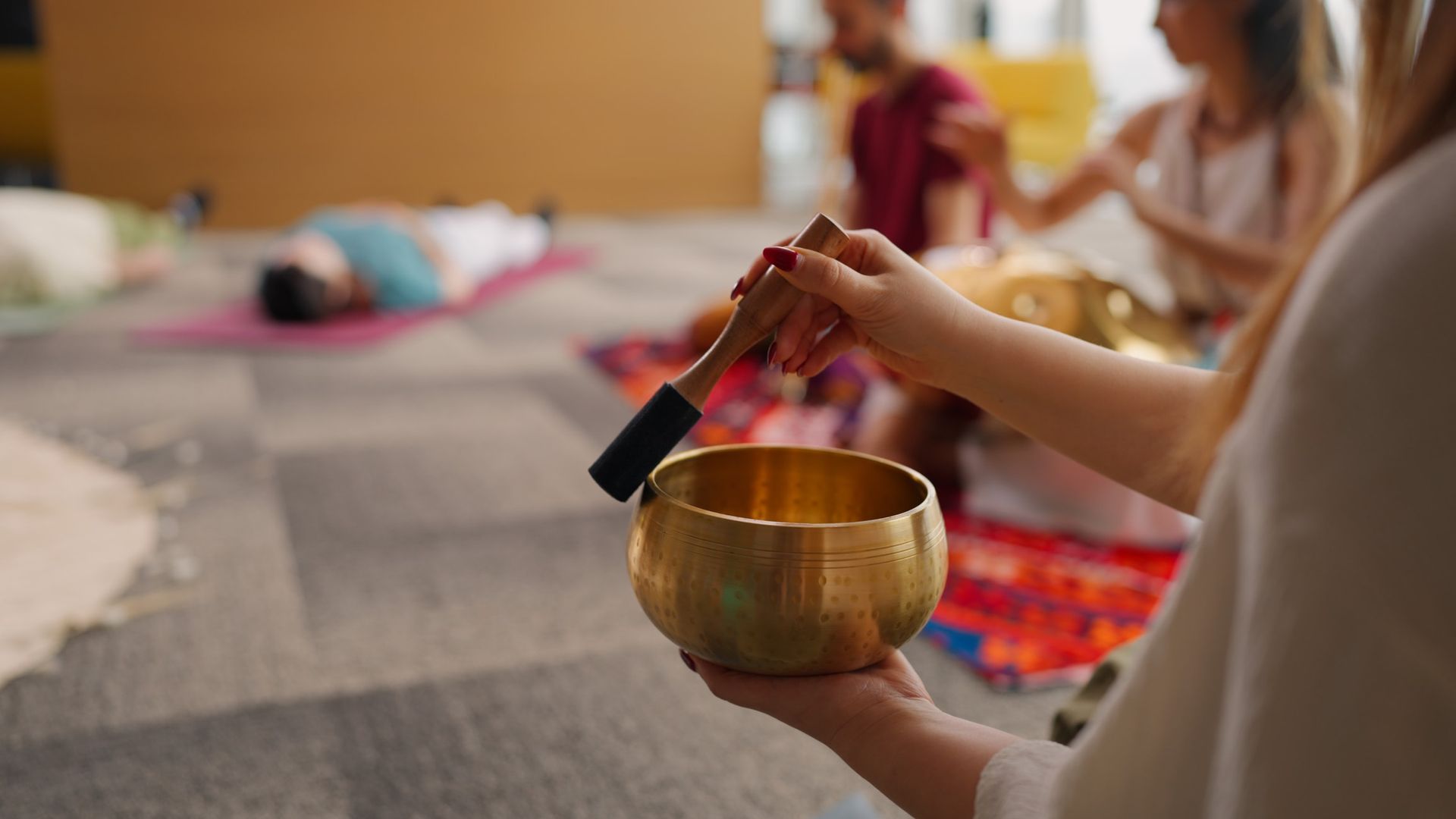 Woman striking a golden singing bowl with a mallet during a sound healing session. Others are visible in the background.