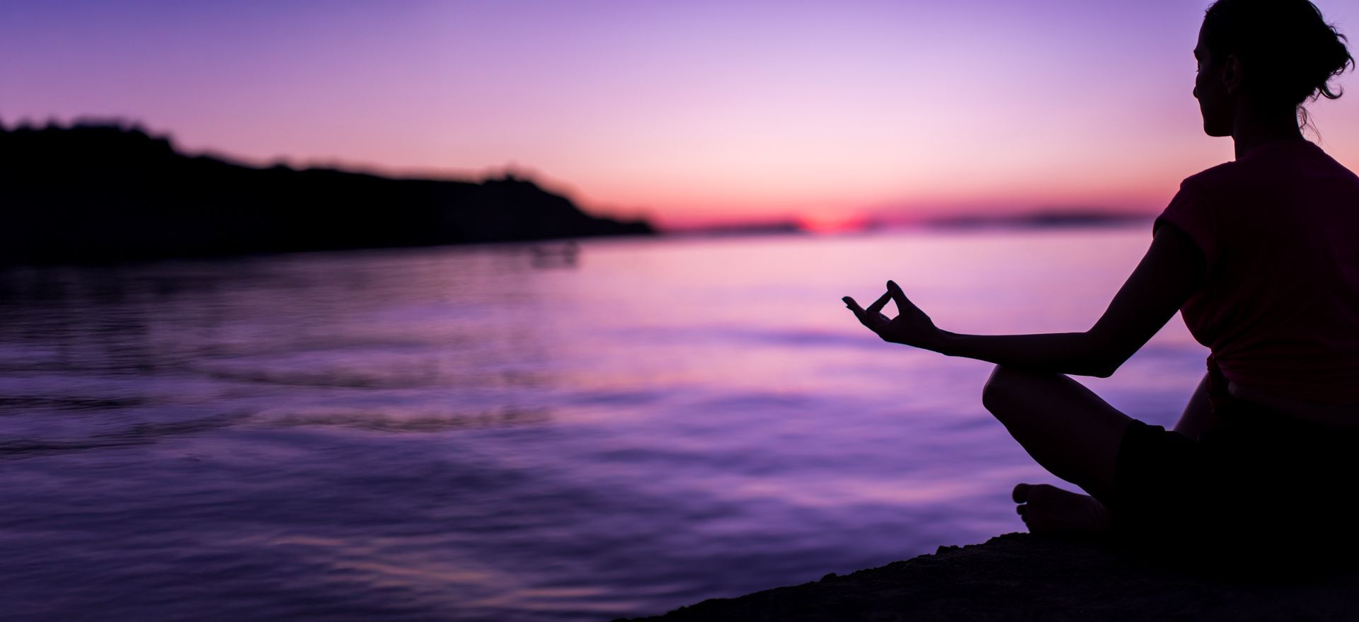 A woman is sitting in a lotus position on the beach at sunset.