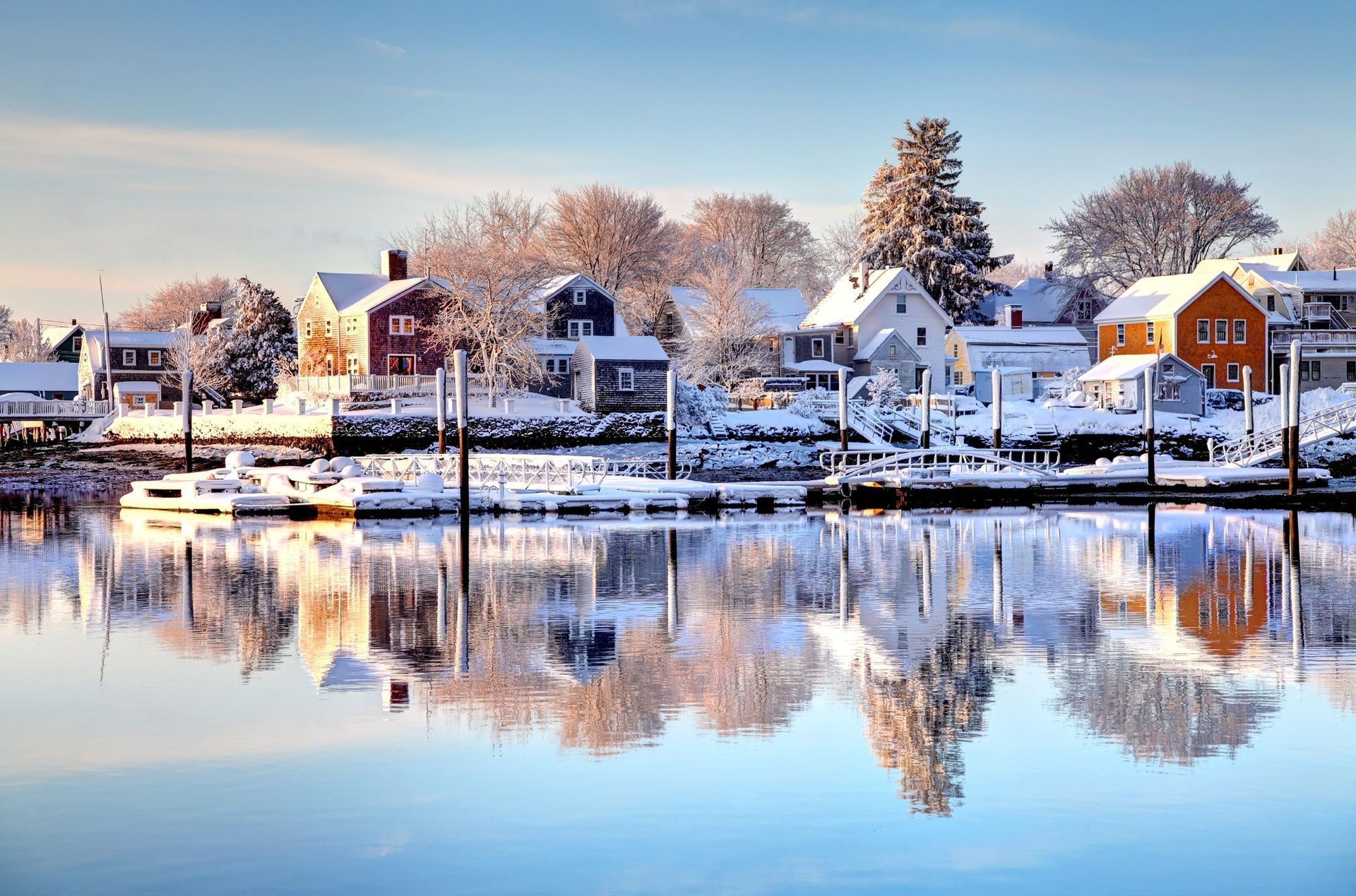 Snow-covered waterfront village with colorful houses reflected in calm water under a blue sky.