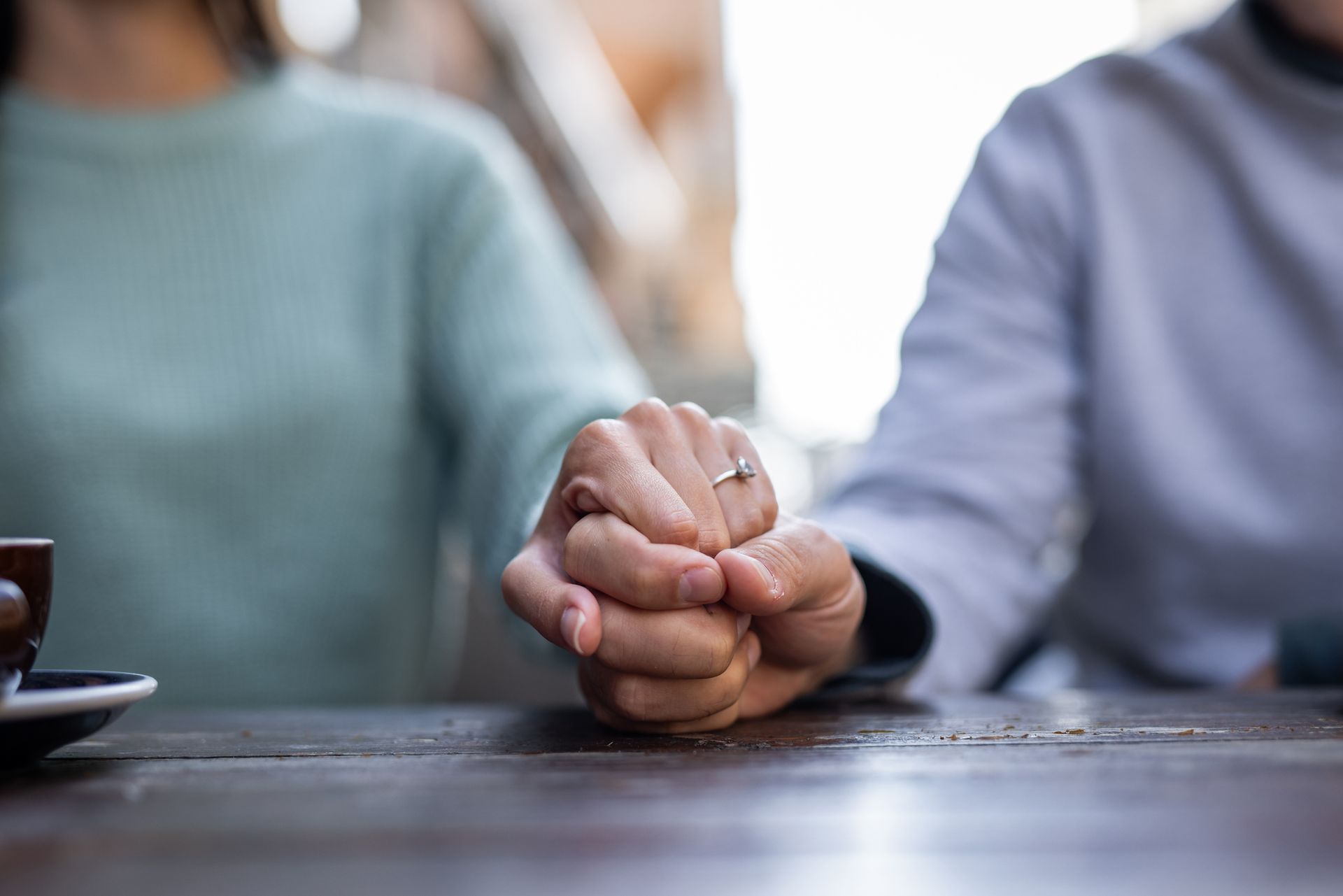 Two people holding hands across a wooden table, one wearing a ring.