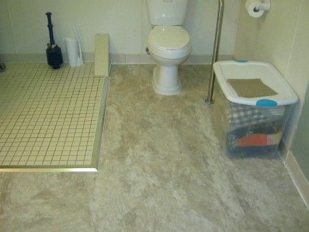 Small bathroom with toilet, tiled shower, floor brush, and plastic storage bin.