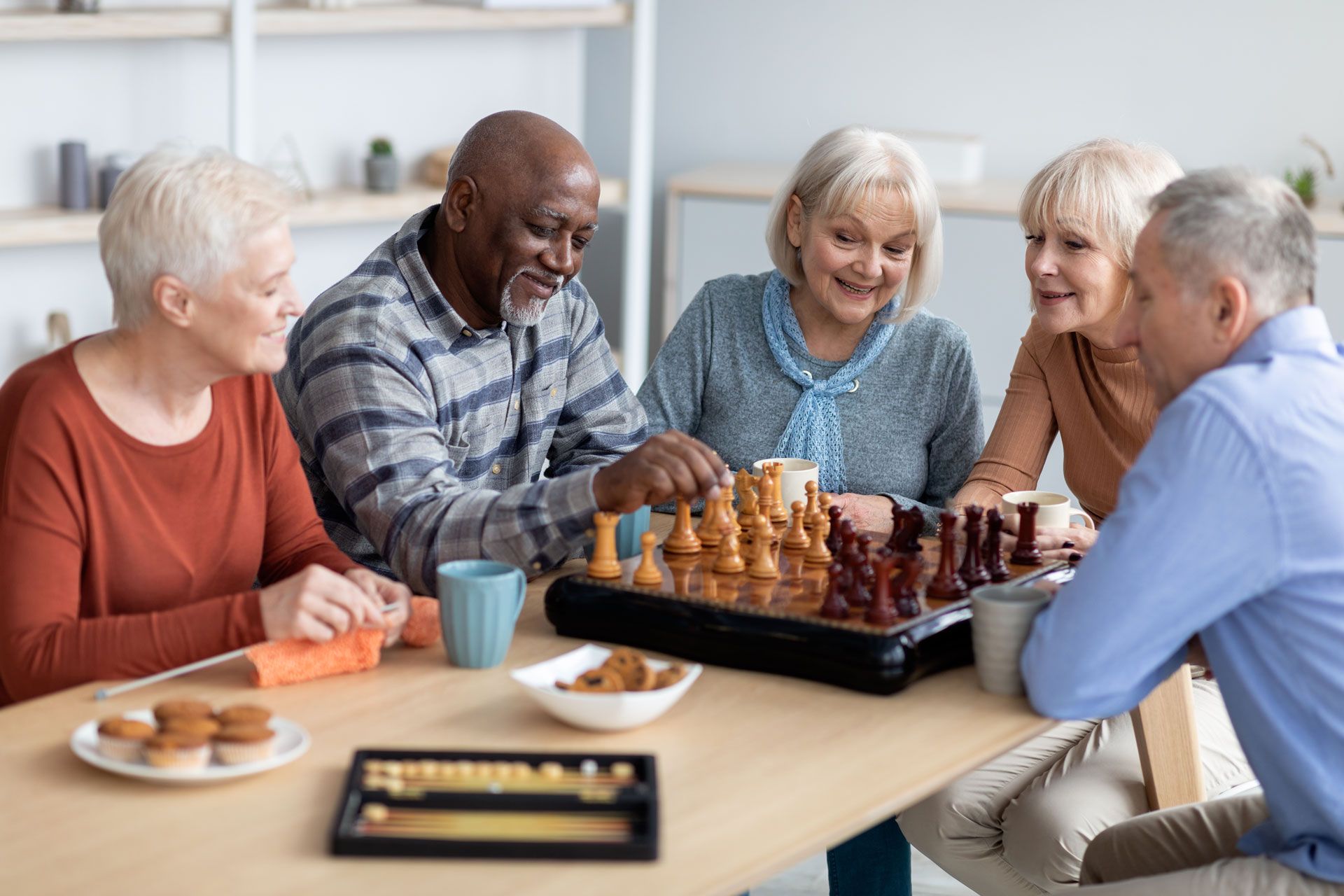 Five people at a table; one is playing chess, others look on. They smile, indoors, cups and snacks present.